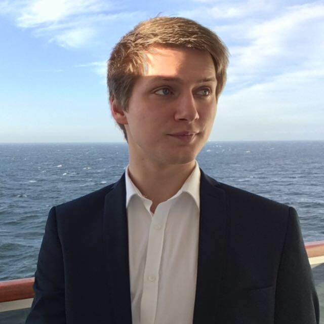 Profile photo of Adam Greenough, he is looking right on a ship, there is the open sea and blue sky in the background